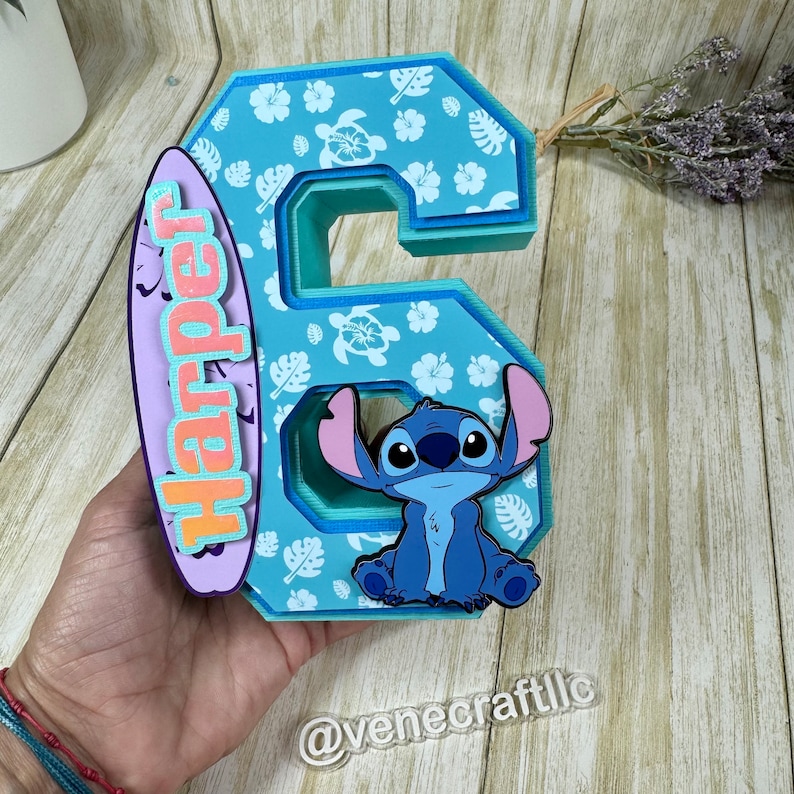 May include: A layered, blue number 6 with a cartoon character and a surfboard. The number is decorated with white floral and turtle patterns. The surfboard has the name "Harper" in orange and pink.