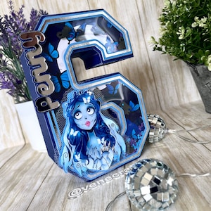 May include: A blue and silver 3D number six with a cutout window featuring a cartoon character with long black hair and a white dress. The character is surrounded by blue butterflies. The number is decorated with silver glitter and has the word "Mama" written on the side.