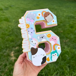 Op de afbeelding: Een decoratief cijfer zes gemaakt van gelaagd papier, met cartoon dierenfiguren, regenbogen en sinaasappelschijfjes. Het cijfer heeft een lichtblauwe voorkant en een roze binnenkant. Een naam staat verticaal op de zijkant.