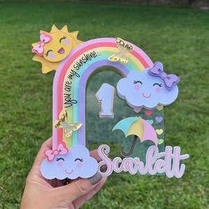 May include: A rainbow-themed first birthday cake topper featuring a sun, clouds, butterflies, and an umbrella. The topper is made from paper and has a clear acrylic base. The text "You are my sunshine..." is written on the rainbow. The number "1" is in the centre of the topper. The name "Scarlett" is written at the bottom of the topper.