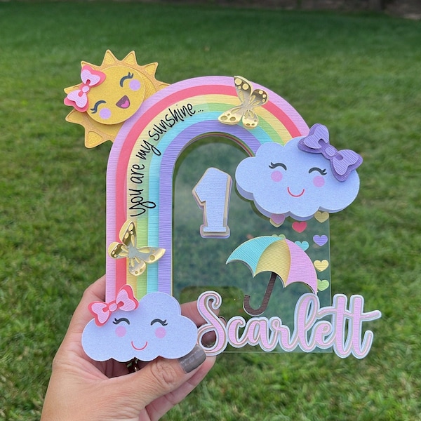 Könnte beinhalten: Ein regenbogenfarbener erster Geburtstagstortenaufsatz mit einer Sonne, Wolken, Schmetterlingen und einem Regenschirm. Der Aufsatz ist aus Papier und hat eine klare Acrylbasis. Der Text "You are my sunshine..." ist auf dem Regenbogen geschrieben. Die Zahl "1" befindet sich in der Mitte des Aufsatzes. Der Name "Scarlett" ist unten auf dem Aufsatz geschrieben.