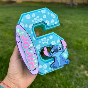 May include: A light blue number 6 with a tropical floral pattern and a cartoon character. A purple surfboard with the name "Harper" is attached to the number. The character is a blue alien with large ears.