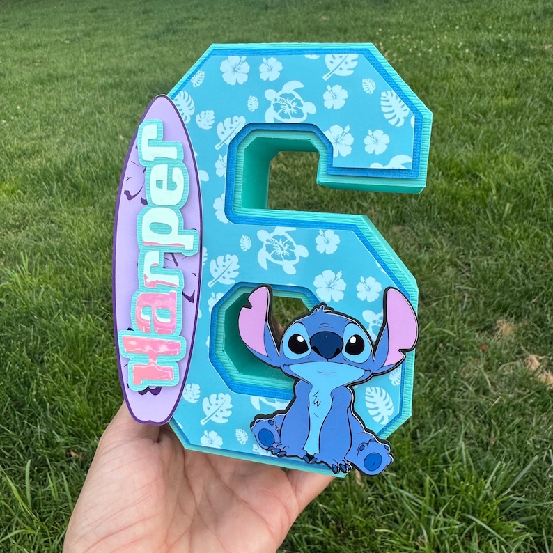 May include: A layered, turquoise number six with a tropical pattern, a cartoon character, and a surfboard with the name "Harper". The number has a 3D effect, with a light blue interior and a dark blue outline. The surfboard is purple and pink.