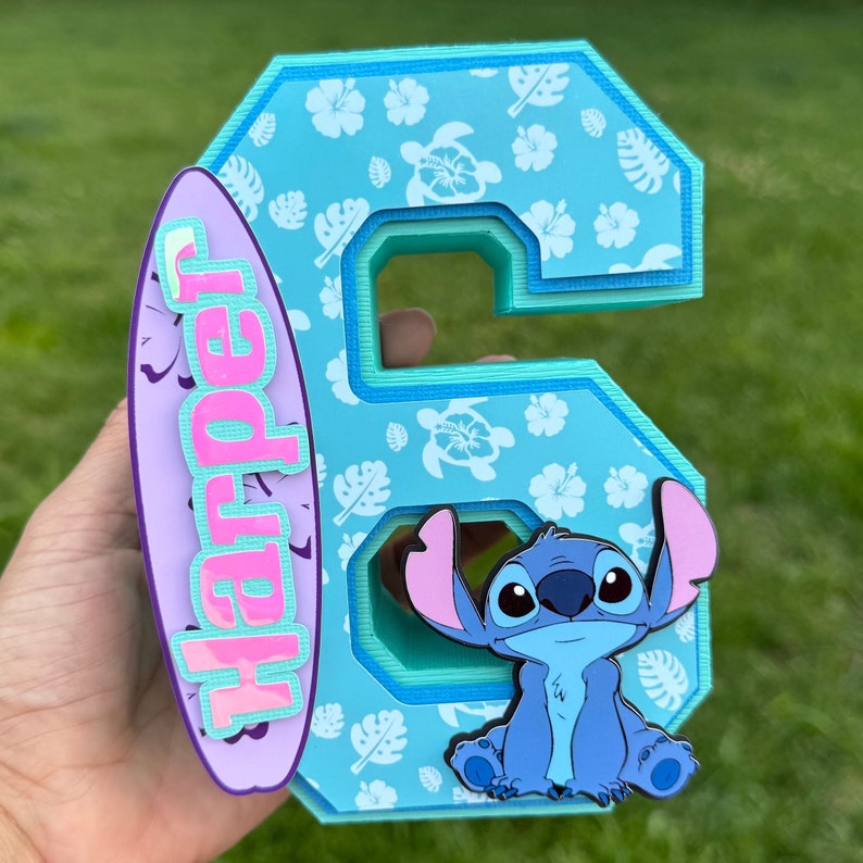 May include: A layered, light blue number six with a tropical floral pattern and a cartoon character. A purple surfboard with the name "Harper" in pink and light blue is attached to the side. The character is a blue alien with large ears.