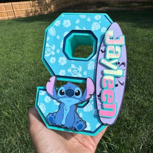 May include: A layered, light blue number nine with a cartoon Stitch character and a surfboard with the name "Jayleen". The number has a floral pattern and a teal interior. The surfboard is purple and pink.