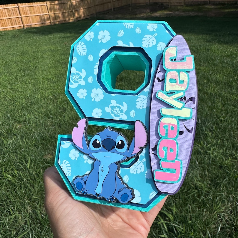 May include: A layered, light blue number nine with a cartoon Stitch character and a surfboard with the name "Jayleen". The number has a floral pattern and a teal interior. The surfboard is purple and pink.