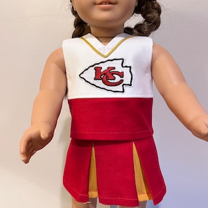 May include: A doll wearing a cheerleader outfit. The top is white with a red and gold logo and trim. The skirt is red with gold pleats. The outfit is designed to resemble a sports team uniform.