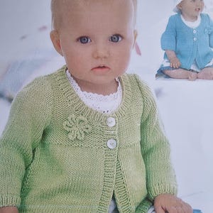 Op de afbeelding: Een groene gebreide cardigan met een bloemetjespatroon op de voorkant en knoopsluiting. De cardigan wordt gedragen door een baby die op een wit oppervlak zit.