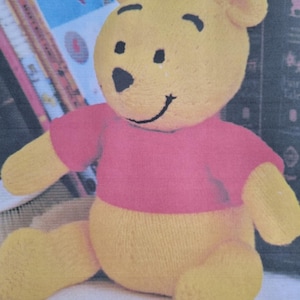 以下が含まれることがあります： 赤いシャツを着たニットのくまのプーさんのぬいぐるみ。くまは本の上に座っています。