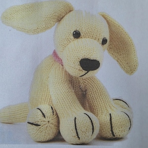 May include: A knitted dog toy in a light yellow color. The dog has large floppy ears, black eyes, and a black nose. It is wearing a pink collar. The paws have black stitched details. The toy is sitting down.