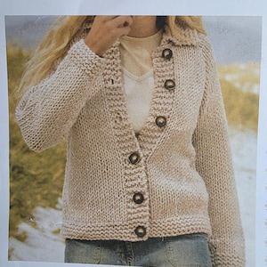Può includere: Un cardigan beige lavorato a maglia con collo e maniche lunghe. Il maglione presenta grandi bottoni rotondi e marroni sul davanti. La maglia ha un aspetto strutturato e un orlo e polsini a coste. L'indumento è mostrato su una persona che indossa una camicia bianca e jeans blu.