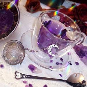 Könnte beinhalten: Eine gläserne Teetasse mit einem violetten Kräutertee und einem silbernen Tee-Infuser mit einem Halbmond-Anhänger. Die Teetasse steht auf einem Untersetzer, umgeben von Amethyst-Kristallen. Ein silberner Teesieb und ein silberner Löffel sind ebenfalls im Bild.
