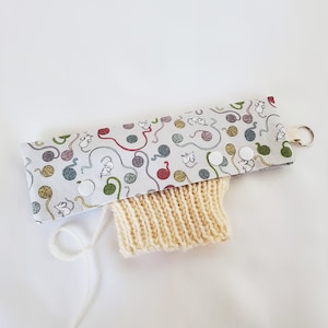 May include: A beige knitted arm warmer with a white fabric cuff featuring a repeating pattern of yarn balls and mice. The cuff has white snap buttons.