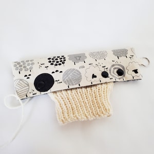 May include: A white knitted cup cosy with a black and white sheep print fabric cover. The fabric has a black button closure.