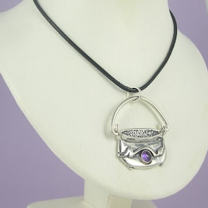 Könnte beinhalten: Eine silberne Halskette mit einem kleinen Kessel-Anhänger und einem violetten Amethyst-Edelstein. Die Halskette ist an einem schwarzen Band.