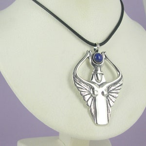 Könnte beinhalten: Eine silberne Anhängerkette mit einer stilisierten Figur mit ausgebreiteten Flügeln und einem zentralen blauen Edelstein. Der Anhänger hängt an einer schwarzen Kordel und zeigt detaillierte Designs und eine polierte Oberfläche. Die Halskette wird auf einer weißen Büste präsentiert.