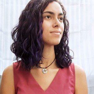 Könnte beinhalten: Eine Frau mit langen, dunkelvioletten Locken trägt ein rotes, ärmelloses Oberteil und eine silberne Halskette mit einem Mond- und Sternanhänger.
