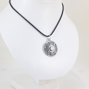 Könnte beinhalten: Eine silberne Halskette mit einem runden Anhänger, der ein Sonnenmotiv mit einem weißen Stein in der Mitte zeigt. Die Halskette ist an einem schwarzen Band.