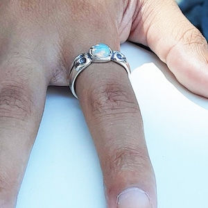 Könnte beinhalten: Ein silberner Ring mit einem weißen Opal-Edelstein und schwarzen Akzenten. Der Ring wird an einem Finger getragen.