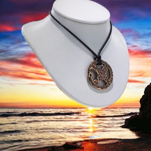 May include: A bronze pendant necklace with a circular design featuring a Celtic knot pattern. The necklace is displayed on a white mannequin against a sunset background.