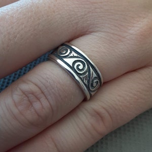 May include: A silver ring with a black swirl design. The ring is worn on a person's finger.