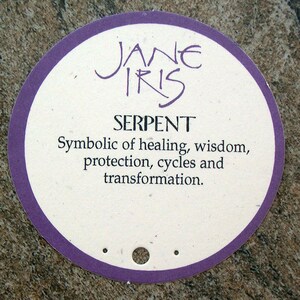 May include: A round, purple-bordered tag with the text "JANE IRIS" and "SERPENT" in black. The tag also includes the text "Symbolic of healing, wisdom, protection, cycles and transformation."