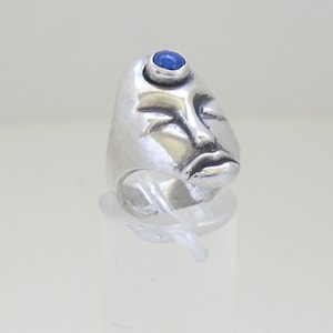 May include: A silver ring with a face design and a blue gemstone in the center of the forehead.