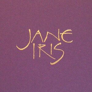 May include: A gold cursive signature that reads "Jane Iris" on a purple background.