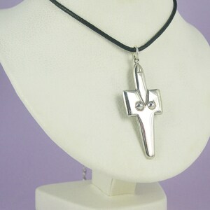 May include: A silver cross pendant with a black cord necklace. The cross has a unique design with two small circles on the front.