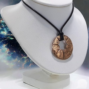 May include: A round, copper-colored pendant with a starburst design hangs from a black leather cord. The pendant is on a white display stand.
