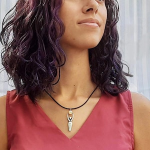 May include: A silver necklace with a pendant featuring a stylized female figure holding a small, round, yellow stone. The necklace is worn by a person with dark purple hair.