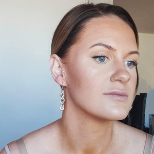 Peut inclure: Une femme aux cheveux châtains clairs et aux yeux bleus porte une boucle d'oreille en argent en forme de nœud infini. La boucle d'oreille pend de son lobe d'oreille gauche.