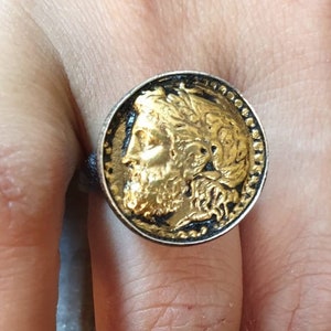 May include: A silver ring with a large, round, gold-colored coin design featuring a profile of a man with a beard and a laurel wreath. The ring is worn on a finger.