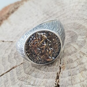 Puede incluir: Un anillo de plata con una cara redonda y texturizada que presenta un dise&ntilde;o detallado en plata oxidada. El anillo est&aacute; sobre una superficie de madera.
