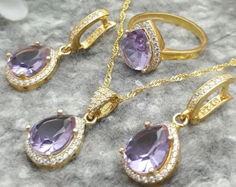 Könnte beinhalten: Ein Gold-Set mit Halskette, Ring und Ohrringen mit einem tropfenförmigen violetten Edelstein, der von klaren Zirkonia-Steinen umgeben ist.
