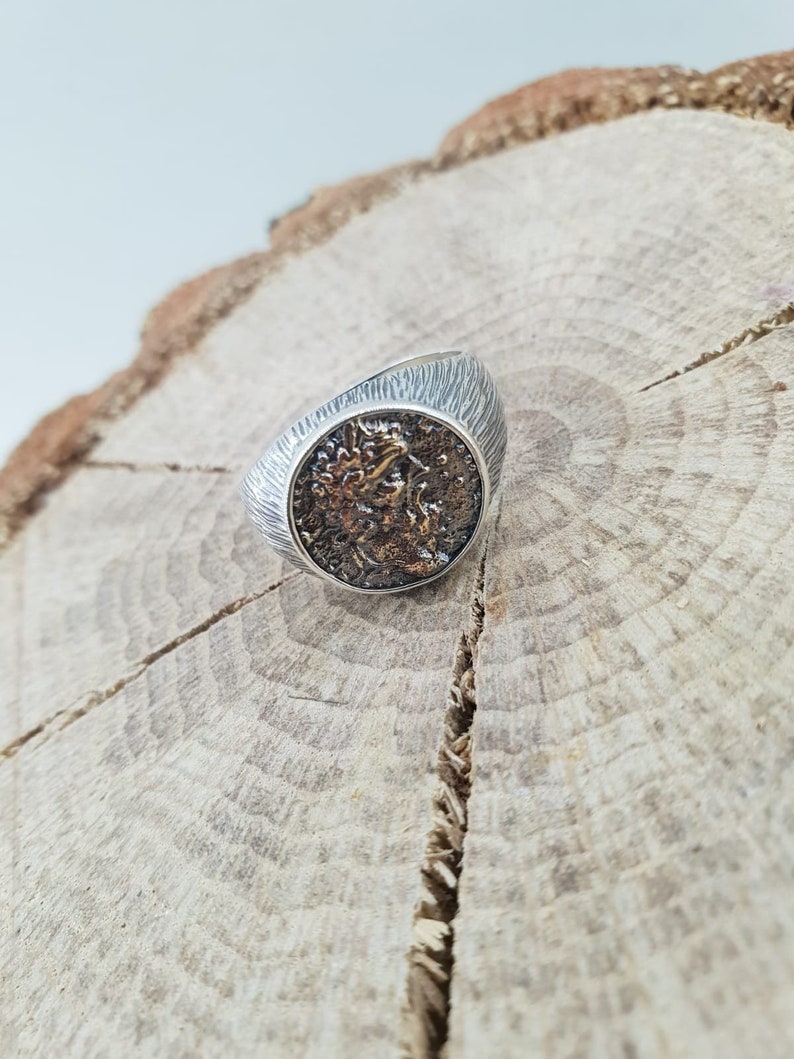 Puede incluir: Un anillo de plata con una cara redonda y texturizada que presenta un dise&ntilde;o detallado en plata oxidada. El anillo est&aacute; sobre una superficie de madera.