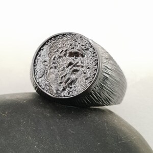 Puede incluir: Un anillo de plata con una banda texturizada y un dise&ntilde;o redondo en relieve en la parte superior. El dise&ntilde;o presenta una imagen detallada y texturizada de una cara.