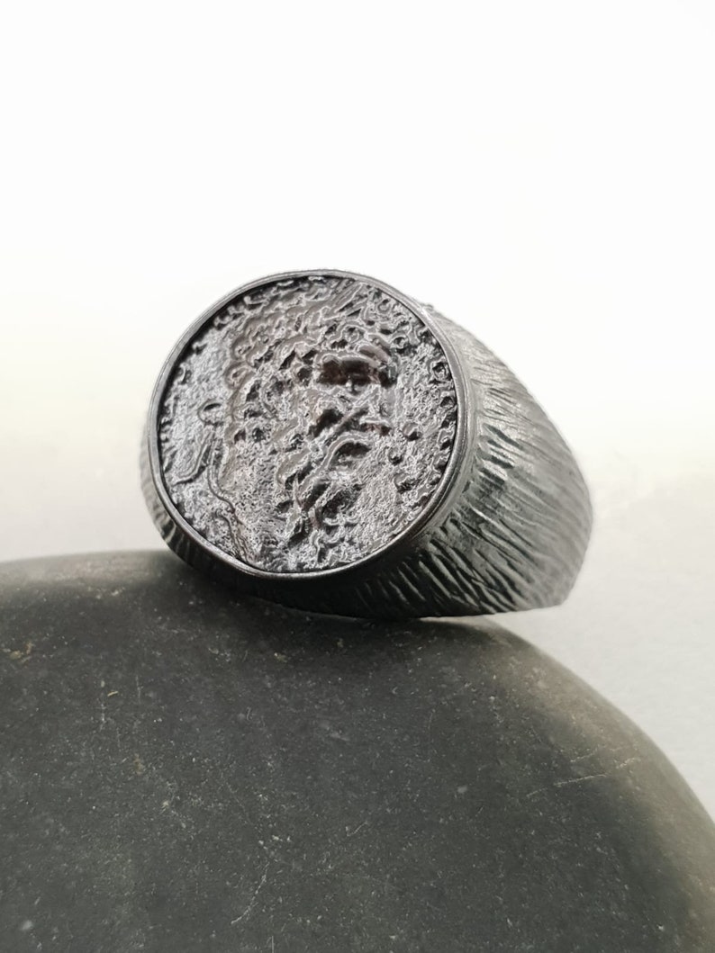Puede incluir: Un anillo de plata con una banda texturizada y un dise&ntilde;o redondo en relieve en la parte superior. El dise&ntilde;o presenta una imagen detallada y texturizada de una cara.