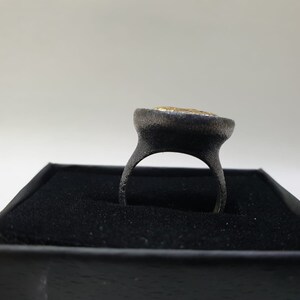 May include: A black ring with a gold-colored stone set in the center. The ring is displayed in a black box.