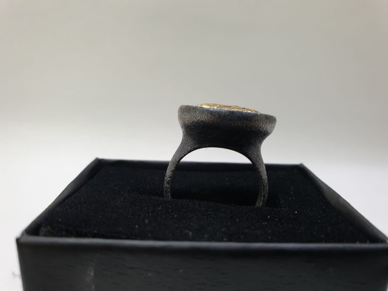 May include: A black ring with a gold-colored stone set in the center. The ring is displayed in a black box.