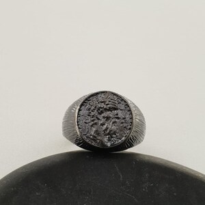 Puede incluir: Un anillo negro con una banda texturizada y un dise&ntilde;o redondo en relieve que presenta un perfil detallado de la cabeza de una persona.