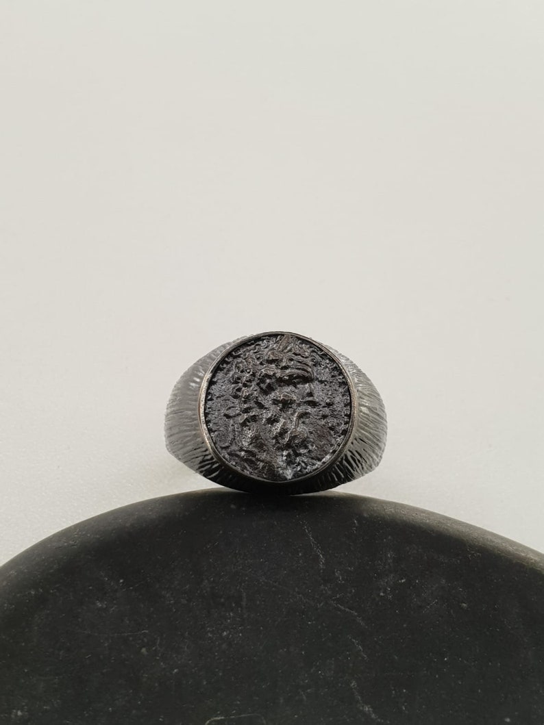 Puede incluir: Un anillo negro con una banda texturizada y un dise&ntilde;o redondo en relieve que presenta un perfil detallado de la cabeza de una persona.