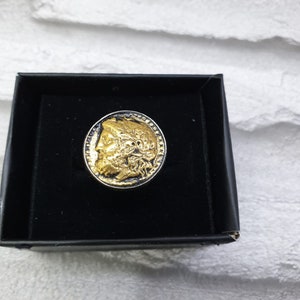 May include: A gold-toned ring with a detailed profile of a person's face on the top. The ring is in a black velvet box.