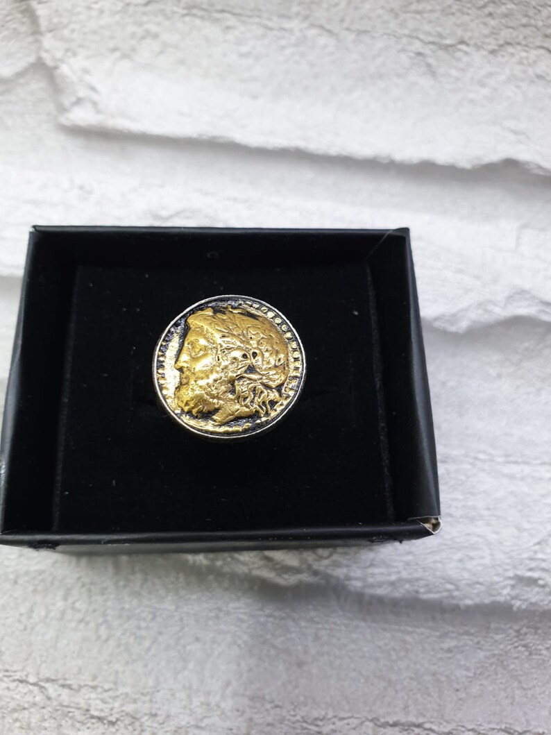May include: A gold-toned ring with a detailed profile of a person's face on the top. The ring is in a black velvet box.