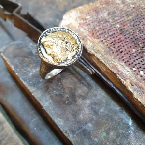 May include: A silver ring with a black and gold coin-shaped top featuring a detailed profile of a person's face.