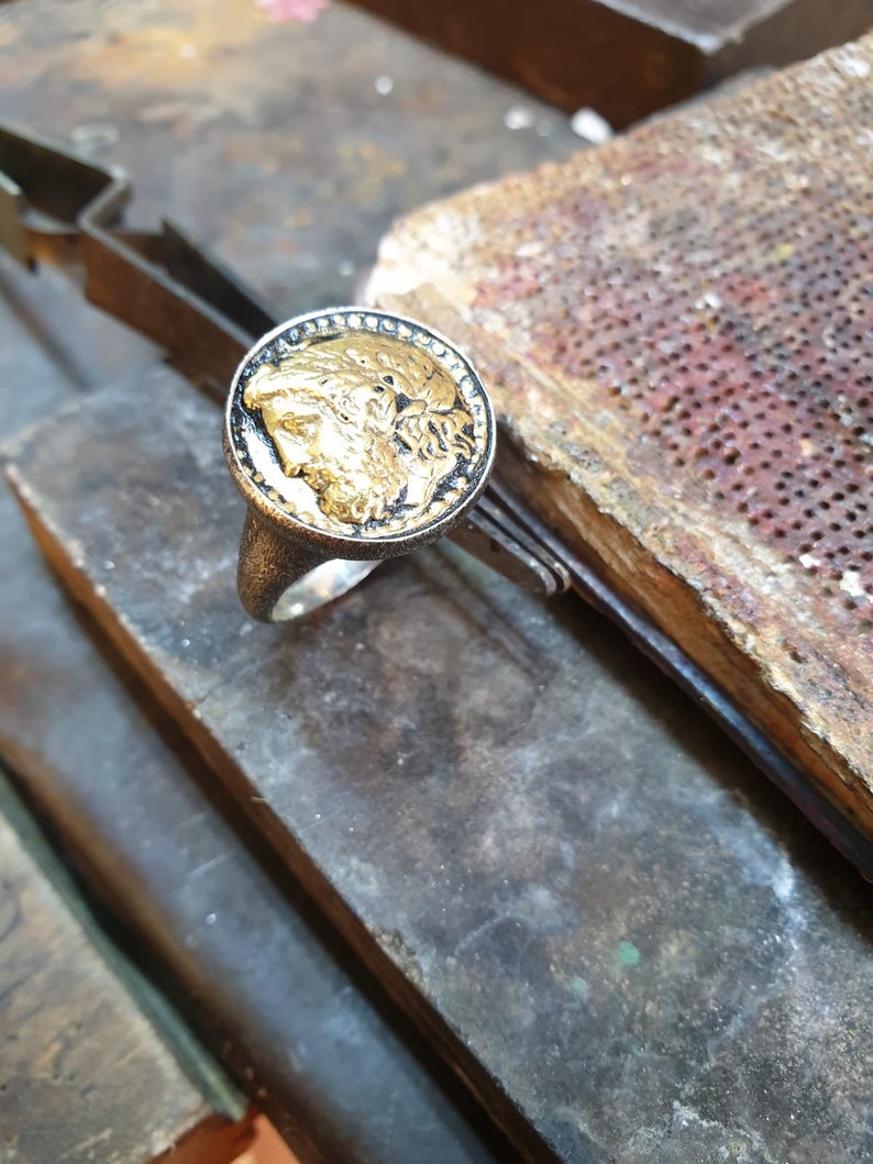 May include: A silver ring with a black and gold coin-shaped top featuring a detailed profile of a person's face.
