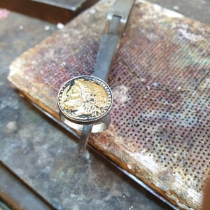 May include: A silver ring with a gold coin design featuring a detailed image of a person's face. The ring is resting on a wooden surface with a perforated metal sheet.