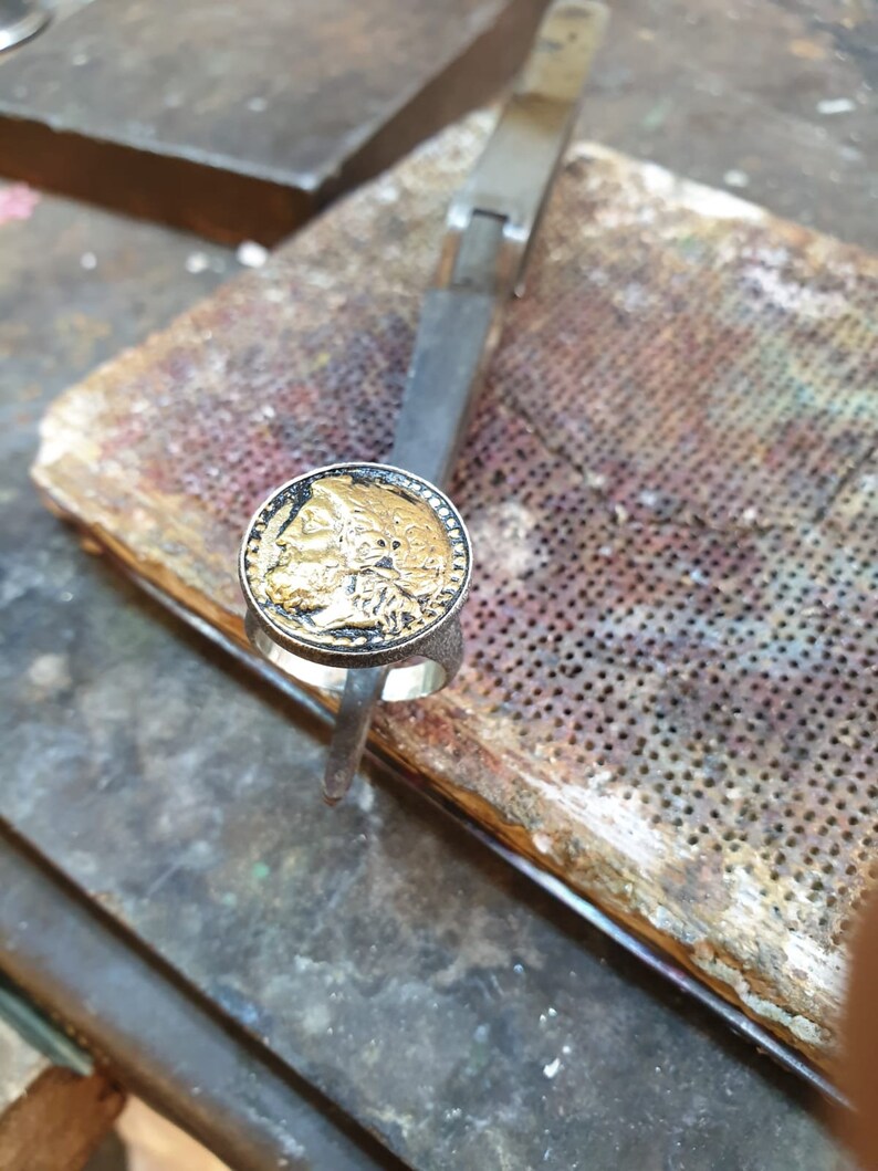 May include: A silver ring with a gold coin design featuring a detailed image of a person's face. The ring is resting on a wooden surface with a perforated metal sheet.