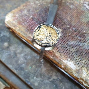 May include: A silver ring with a gold-toned round face featuring a detailed profile of a person's face.
