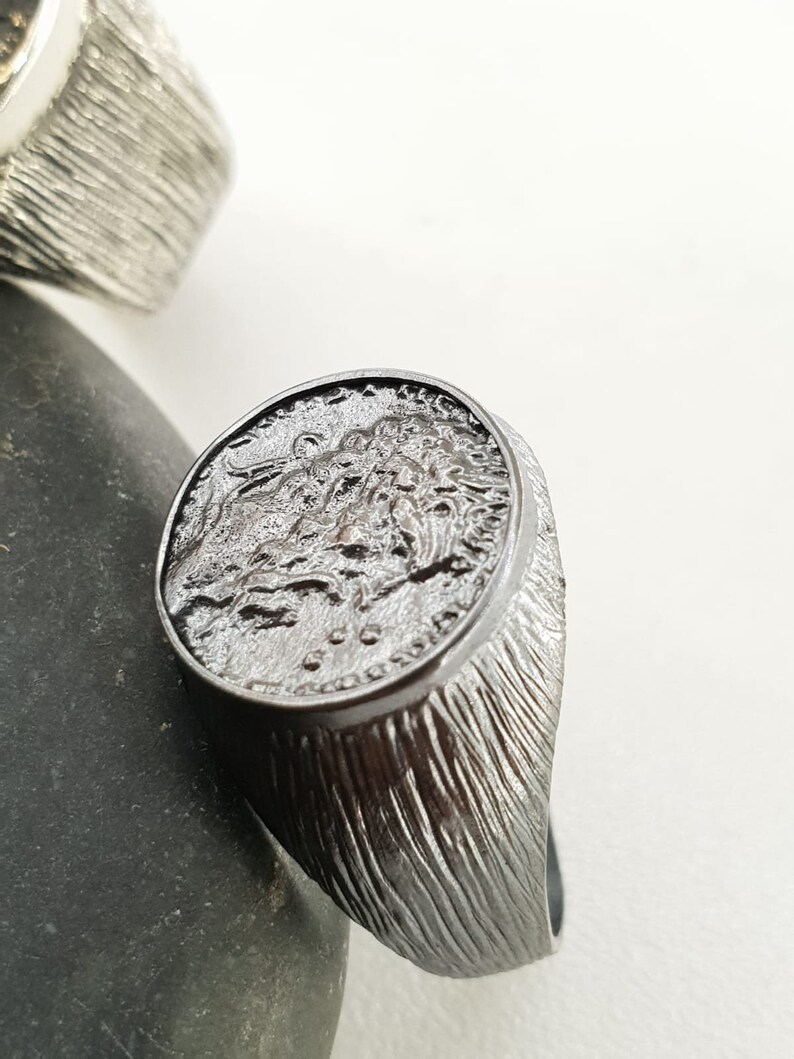 Puede incluir: Un anillo de plata con una banda texturizada y un dise&ntilde;o redondo y detallado en la parte superior. El dise&ntilde;o presenta una imagen estilizada de una cabeza de le&oacute;n.
